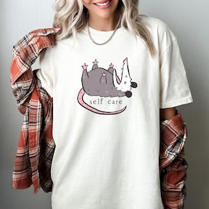 May include: White t-shirt with a cartoon possum illustration and the text "self care".
