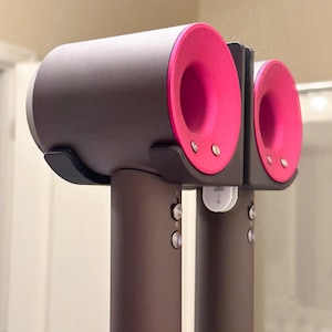 May include: A black hair dryer with two pink attachments. The dryer has two buttons on the side and a white label with an arrow pointing down.