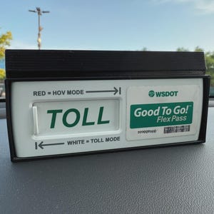 May include: A black and white electronic toll transponder with the word "TOLL" in green. The device has a "Good To Go! Flex Pass" sticker and is labeled "RED = HOV MODE" and "WHITE = TOLL MODE".