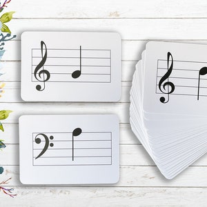 May include: A set of white flashcards with musical notes and clefs printed on them. The flashcards are stacked on a white wooden surface.