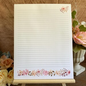 May include: A rectangular sheet of white stationery paper featuring horizontal black lines for writing. The paper is decorated with a small floral cluster in the top right corner and a wide floral border along the bottom edge.