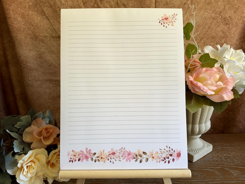 May include: A rectangular sheet of white stationery paper featuring horizontal black lines for writing. The paper is decorated with a small floral cluster in the top right corner and a wide floral border along the bottom edge.