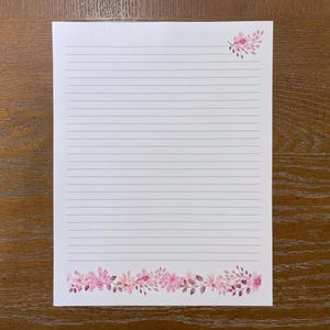 May include: A white sheet of lined stationery paper featuring a horizontal border of pink watercolor flowers and leaves at the bottom edge and a smaller matching floral cluster in the top right corner of the page.