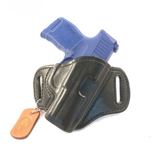 May include: A black leather holster with a blue handgun inside. The holster has a belt loop and a small brown leather tag attached to it.