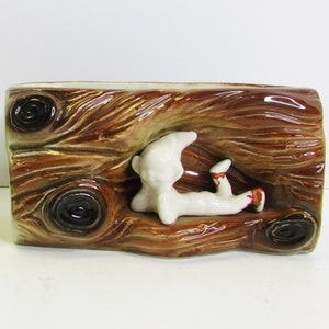 May include: A ceramic planter shaped like a log with a white figurine of a person sitting on top. The planter is brown with a wood grain pattern.