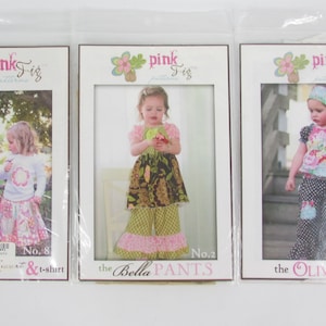May include: Three sewing patterns for children's clothing. The patterns are for a t-shirt, pants, and a skirt. Each pattern features a different design and is packaged in a clear plastic bag. The patterns are from Pink Fig Patterns.