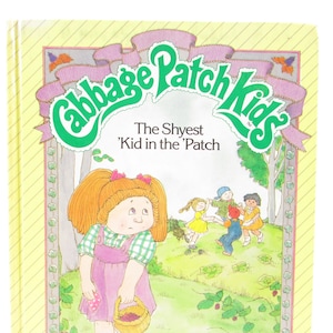 May include: A yellow book cover with the title "Cabbage Patch Kids" in green letters. The book features a picture of a Cabbage Patch Kid doll with red hair and a pink dress, holding a basket of strawberries. The text "The Shyest 'Kid in the 'Patch" is printed below the title.