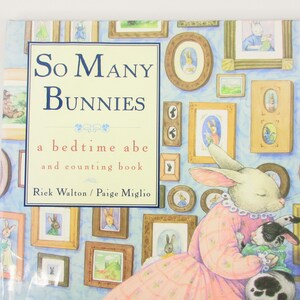 May include: A children's book titled "So Many Bunnies: a bedtime abe and counting book" by Rick Walton and Paige Miglio. The cover features a white bunny in a pink dress holding a black and white bunny. The background is a light blue wall with framed pictures of bunnies.