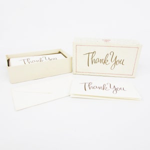 May include: A box of thank you cards with a white background and brown cursive writing. The box has a pink and white patterned design with the words "Thank You" in brown cursive writing.