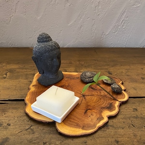 Könnte beinhalten: Ein dekoratives Holztablett mit natürlicher Kante, mit einem kleinen grauen Buddha-Kopf, einem weißen quadratischen Kerzenhalter, kleinen Steinen und einem Zweig Grün. Das Tablett steht auf einer rustikalen Holzoberfläche.
