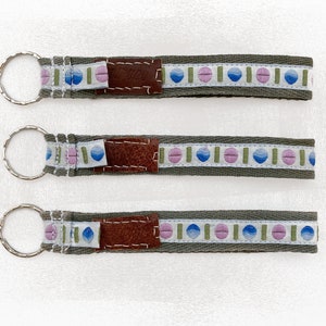 Könnte beinhalten: Drei Schlüsselanhänger mit olivgrünem Stoff und einem gemusterten weißen Band mit blauen, rosa und grünen Formen. Jeder Schlüsselanhänger hat einen braunen Lederakzent und einen silbernen Schlüsselring.