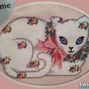 May include: Cross-stitch design of a white cat with blue eyes, a red bow, and floral accents. The cat is surrounded by a pink oval border. The design is a vintage craft project.