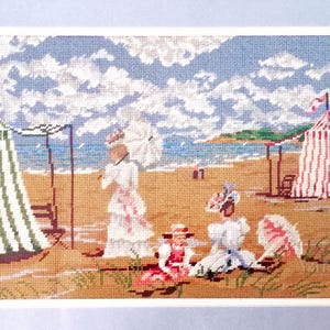 May include: A needlepoint scene of a beach with figures in period clothing, parasols, and striped tents. The sky is blue with white clouds, and the sand is tan. The artwork is framed.