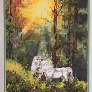 May include: Cross-stitch artwork depicting two white unicorns in a forest clearing. The scene is bathed in sunlight filtering through the trees, with a vibrant mix of green, yellow, and brown hues. The text "Variety Ops" is visible.