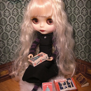 K&ouml;nnte beinhalten: Eine Puppe mit langen blonden Haaren h&auml;lt eine Schachtel mit Vintage-Weihnachtsbaumlichtern der Marke Shiny Brite. Die Puppe tr&auml;gt ein schwarzes Kleid mit wei&szlig;em Kragen und sitzt auf einer flauschigen wei&szlig;en Oberfl&auml;che. Die Puppe ist umgeben von Schachteln mit Vintage-Weihnachtsbaumlichtern, darunter Noma Lites Candoler, Noma 25-Light Outdoor Waterproof und Noma Is Feet.