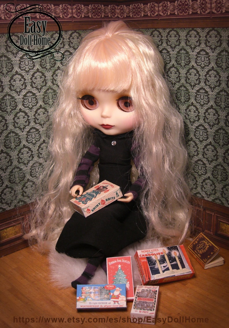 K&ouml;nnte beinhalten: Eine Puppe mit langen blonden Haaren h&auml;lt eine Schachtel mit Vintage-Weihnachtsbaumlichtern der Marke Shiny Brite. Die Puppe tr&auml;gt ein schwarzes Kleid mit wei&szlig;em Kragen und sitzt auf einer flauschigen wei&szlig;en Oberfl&auml;che. Die Puppe ist umgeben von Schachteln mit Vintage-Weihnachtsbaumlichtern, darunter Noma Lites Candoler, Noma 25-Light Outdoor Waterproof und Noma Is Feet.