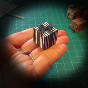 May include: A miniature gift box with black and white vertical stripes and a matching bow. The small present is held in a hand, with other miniature gifts in the background. The box is a cube shape.