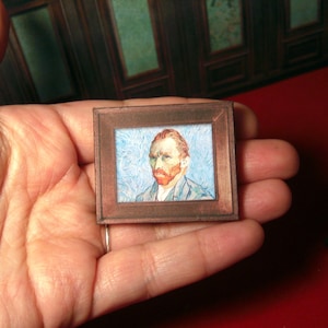 May include: Miniature framed artwork featuring a portrait of Vincent van Gogh. The artwork depicts the artist with a red beard and hair, set against a blue background with swirling brushstrokes. The frame is a dark brown color.