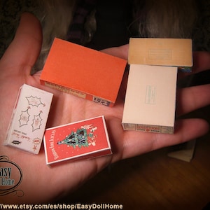 K&ouml;nnte beinhalten: Eine Hand h&auml;lt f&uuml;nf Miniaturboxen. Die Boxen sind mit weihnachtlichen Designs verziert, darunter ein Weihnachtsbaum, Stechpalme und der Text "Ribbon Foil Icicles".