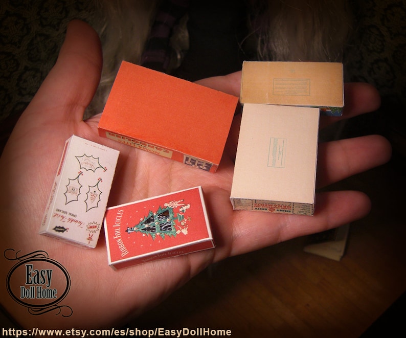 K&ouml;nnte beinhalten: Eine Hand h&auml;lt f&uuml;nf Miniaturboxen. Die Boxen sind mit weihnachtlichen Designs verziert, darunter ein Weihnachtsbaum, Stechpalme und der Text "Ribbon Foil Icicles".