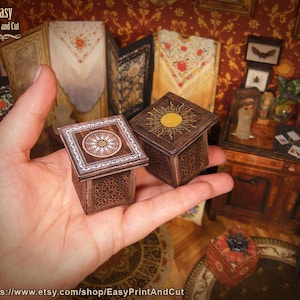 Könnte beinhalten: Zwei Miniatur-Holzkisten mit aufwendigen Designs werden in einer Hand gehalten. Eine Box zeigt ein Sonnendesign, die andere ein kreisförmiges Muster. Die Kisten sind braun mit detaillierten Schnitzereien. Der Hintergrund umfasst Vintage-Dekor und den Text "Easy Print and Cut".