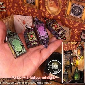 May include: A hand holds several miniature paper lanterns in green, purple, and black, with intricate geometric patterns and a small light. The lanterns are part of a craft project, with the text "Easy Print and Cut" visible.