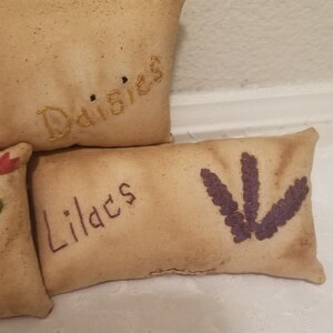 Puede incluir: Tres almohadas peque&ntilde;as y rectangulares con dise&ntilde;os florales bordados. La almohada superior es beige con la palabra "Margaritas" bordada en hilo dorado. La almohada del medio es beige con la palabra "Lirios" bordada en hilo dorado. La almohada inferior es beige con una flor bordada en morado.