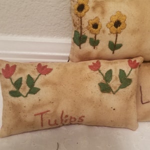 Puede incluir: Dos almohadas de tiro bordadas con dise&ntilde;os florales. La almohada superior tiene tres girasoles amarillos con tallos y hojas verdes. La almohada inferior tiene dos tulipanes rojos con tallos y hojas verdes. La almohada inferior tiene la palabra "Tulips" bordada en rojo.