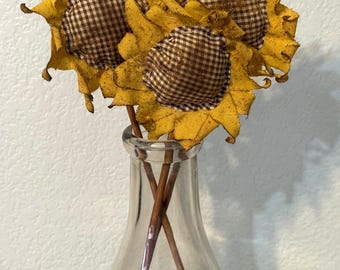 May include: Three fabric sunflowers with mustard yellow petals and brown and white checkered centers, each attached to a thin brown stem. The flowers are arranged inside a clear glass bottle that contains a small amount of dried moss at the base.