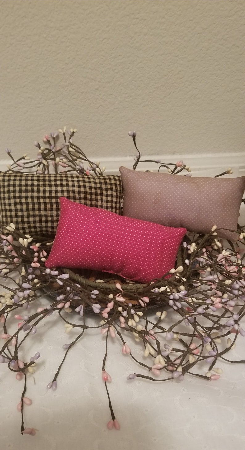 Puede incluir: Tres almohadas decorativas con diferentes patrones. Las almohadas est&aacute;n dispuestas en una cesta de mimbre marr&oacute;n con una corona de ramas de bayas blancas y moradas.