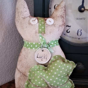May include: A white fabric cat with green polka dot fabric around its neck and a green shamrock with white polka dots. The cat is holding a silver tag that says "Lucky" in black lettering.