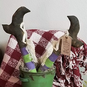 May include: A pair of witch legs with green and purple stripes are sticking out of a green metal bucket. The bucket is sitting on a wooden table with a red and white checkered blanket draped over it. The blanket has fringe on the edges. A small tag with the text "Little Flet" is attached to the legs.