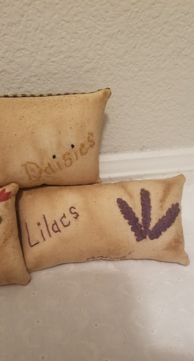 Puede incluir: Tres almohadas peque&ntilde;as y rectangulares con dise&ntilde;os florales bordados. La almohada superior es beige con la palabra "Margaritas" bordada en hilo dorado. La almohada del medio es beige con la palabra "Lirios" bordada en hilo dorado. La almohada inferior es beige con una flor bordada en morado.