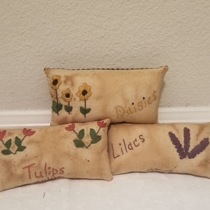 Puede incluir: Cuatro peque&ntilde;os cojines rectangulares de tela beige con flores bordadas y las palabras "Tulips", "Lilacs", "Daisies" y "Daisies".