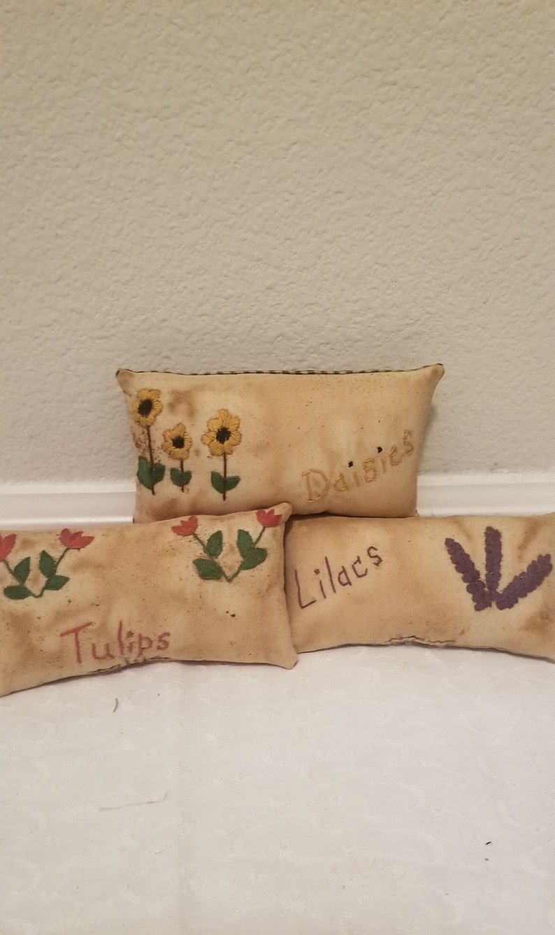 Puede incluir: Cuatro peque&ntilde;os cojines rectangulares de tela beige con flores bordadas y las palabras "Tulips", "Lilacs", "Daisies" y "Daisies".