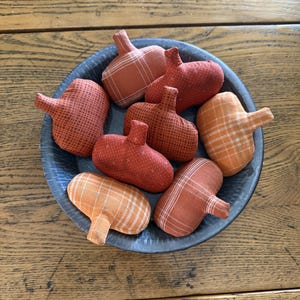 May include: A collection of fabric pumpkins in shades of orange and red, with various patterns including plaid and polka dots, are arranged in a blue ceramic bowl. The pumpkins have small, brown stems.