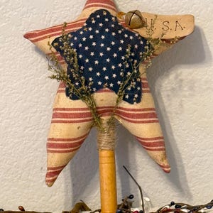 May include: A handmade star-shaped tree topper with a red, white, and blue striped fabric and a blue star with white stars on it. The topper has a wooden dowel and a small bell with a tag that says "U.S.A."