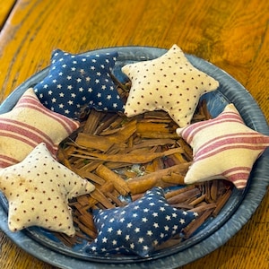 May include: A blue metal pie plate filled with cinnamon sticks and five fabric star-shaped pillows. The pillows are decorated with red and white stripes, blue and white stars, and tan and white stars.