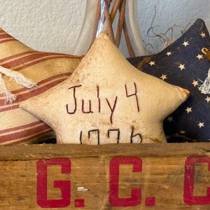 May include: Three star-shaped fabric pillows in a wooden crate. The middle pillow is beige and has the text "July 4 1776" embroidered on it. The crate has the letters "G.C.C." painted in red on the front.