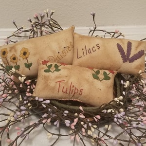 Puede incluir: Cuatro peque&ntilde;os almohadones rectangulares de tela beige con flores bordadas y las palabras "Margaritas", "Lirios", "Tulipanes" y una flor morada. Los almohadones est&aacute;n dispuestos en una cesta con una corona de ramas de bayas blancas y moradas.