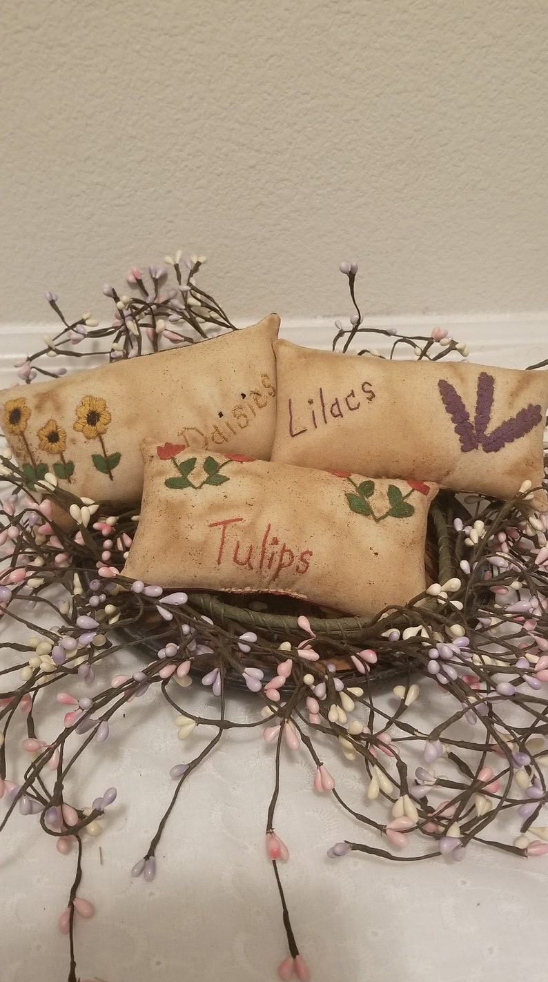 Puede incluir: Cuatro peque&ntilde;os almohadones rectangulares de tela beige con flores bordadas y las palabras "Margaritas", "Lirios", "Tulipanes" y una flor morada. Los almohadones est&aacute;n dispuestos en una cesta con una corona de ramas de bayas blancas y moradas.