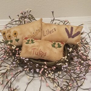 Puede incluir: Cuatro peque&ntilde;os almohadones rectangulares de tela beige con flores bordadas y las palabras "Margaritas", "Lirios", "Tulipanes" y una flor morada. Los almohadones est&aacute;n dispuestos en una cesta forrada con una guirnalda de peque&ntilde;as flores blancas y moradas.