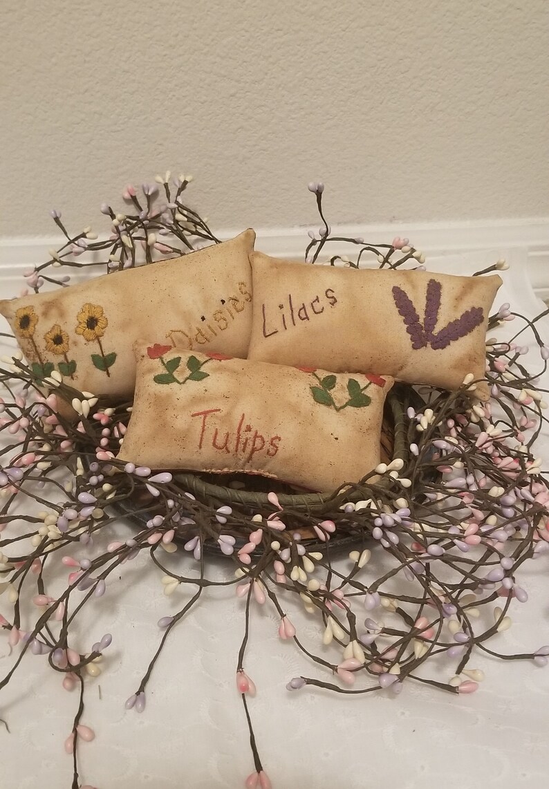 Puede incluir: Cuatro peque&ntilde;os almohadones rectangulares de tela beige con flores bordadas y las palabras "Margaritas", "Lirios", "Tulipanes" y una flor morada. Los almohadones est&aacute;n dispuestos en una cesta forrada con una guirnalda de peque&ntilde;as flores blancas y moradas.