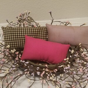 Puede incluir: Tres almohadas decorativas en una cesta de mimbre marr&oacute;n con una corona de ramas de bayas blancas y moradas. Las almohadas son de cuadros blancos y negros, rosa con lunares blancos y morado claro con un patr&oacute;n sutil.