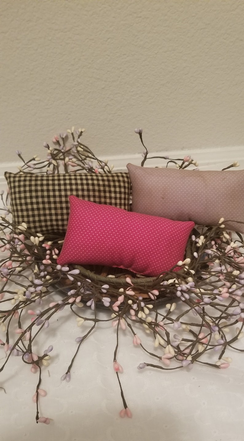 Puede incluir: Tres almohadas decorativas en una cesta de mimbre marr&oacute;n con una corona de ramas de bayas blancas y moradas. Las almohadas son de cuadros blancos y negros, rosa con lunares blancos y morado claro con un patr&oacute;n sutil.