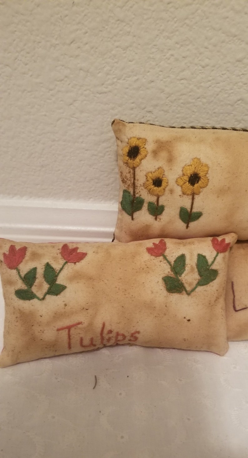 Puede incluir: Dos almohadas de tiro bordadas con dise&ntilde;os florales. La almohada superior tiene tres girasoles amarillos con tallos y hojas verdes. La almohada inferior tiene dos tulipanes rojos con tallos y hojas verdes. La almohada inferior tiene la palabra "Tulips" bordada en rojo.