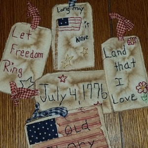 May include: Five fabric tags with patriotic themes. The tags are decorated with red, white, and blue stars, stripes, and flowers. The text on the tags reads: "Let Freedom Ring", "Long may it Wave", "Land that I Love", "July 4, 1776", and "Old Glory".