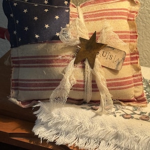 May include: A decorative pillow with a patriotic theme. The pillow features a blue section with white stars and red and white stripes, resembling the American flag. A rusty star and a tag that says "USA" are attached with a white ribbon. The pillow is placed on a fringed white cloth.