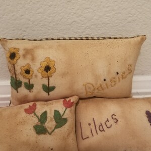 Puede incluir: Tres almohadas de tela bordadas con las palabras "Margaritas", "Lirios" y "Tulipanes" en ellas. Las almohadas son beige con costuras marrones y presentan flores bordadas en amarillo, rojo y verde.