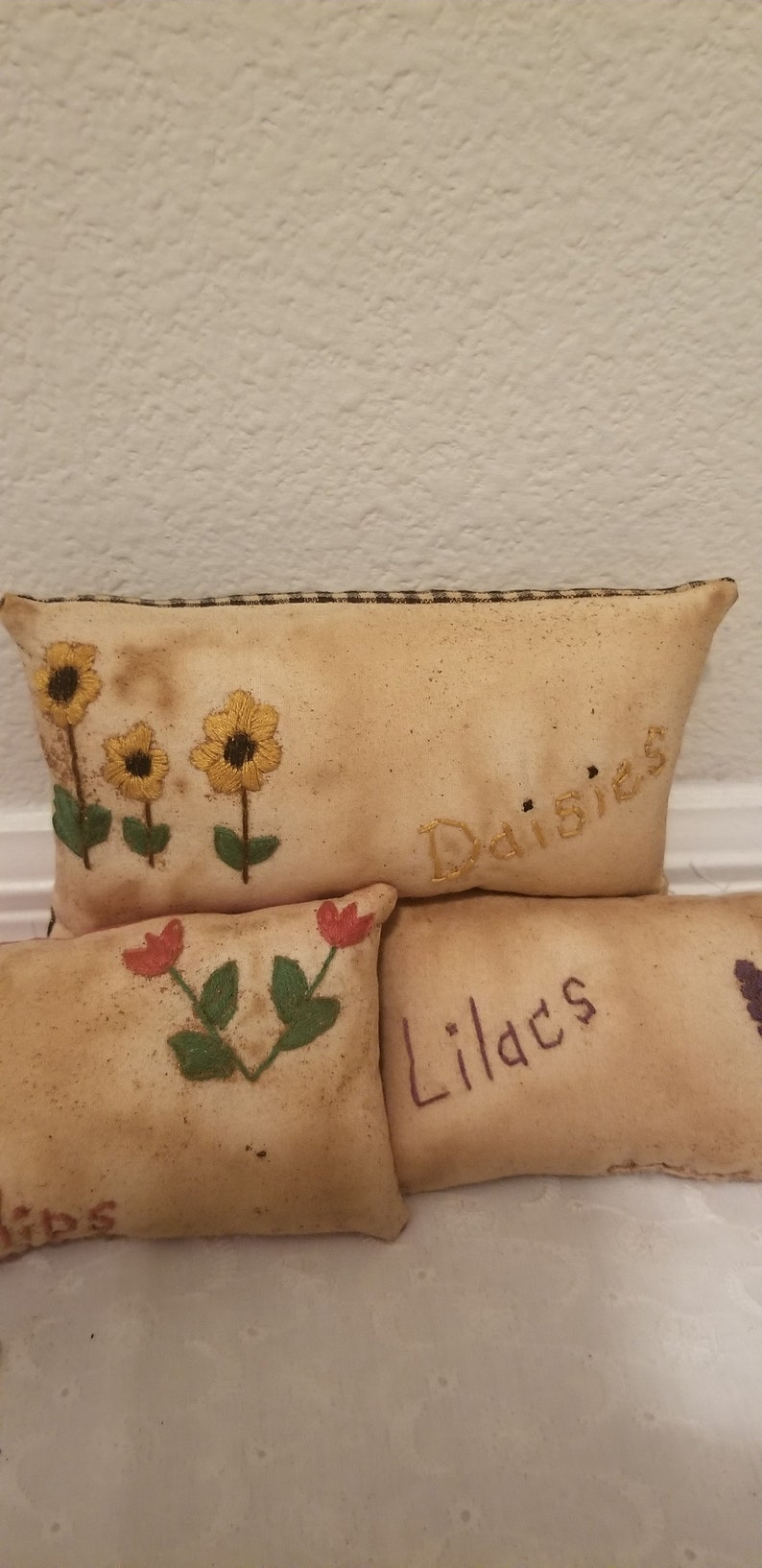 Puede incluir: Tres almohadas de tela bordadas con las palabras "Margaritas", "Lirios" y "Tulipanes" en ellas. Las almohadas son beige con costuras marrones y presentan flores bordadas en amarillo, rojo y verde.